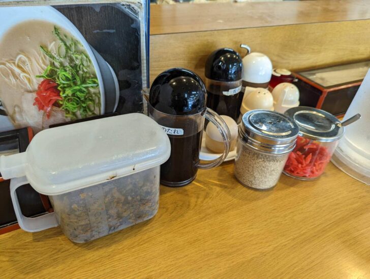 On the table are spicy pickled mustard greens, ramen sauce, ground sesame, pickled ginger, garlic, etc.